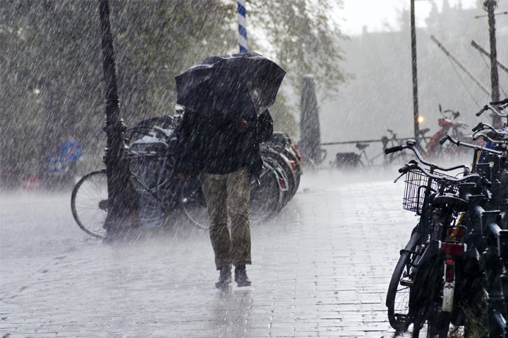 Kuvassa ihminen kävelee kaupunkimaisessa ympäristössä rankkasateessa sateenvarjo suojanaan, tummissa vaatteissa.