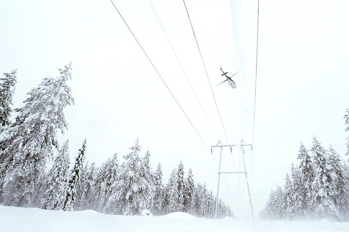 Helikopteri lentaa lumisessa metsämaisemassa voimajohtolinja yllä